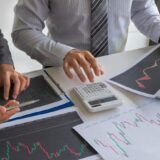 Two business professionals review financial charts: printed line charts, a calculator, and a laptop on a desk |in a meeting setting|, concise and informative for finance context.
