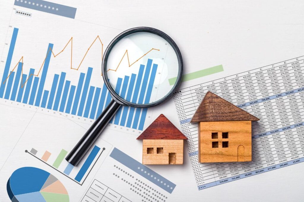 Magnifying glass over financial charts and two wooden house models, implying real estate market analysis