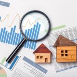 Magnifying glass over financial charts and two wooden house models, implying real estate market analysis
