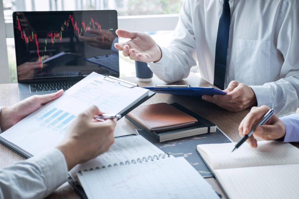 Professionals in a meeting reviewing stock charts on a laptop with notes and reports on the table