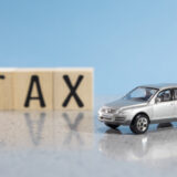 Silver toy car in the foreground with wooden blocks spelling 'TAX' in the background on a blue surface.
