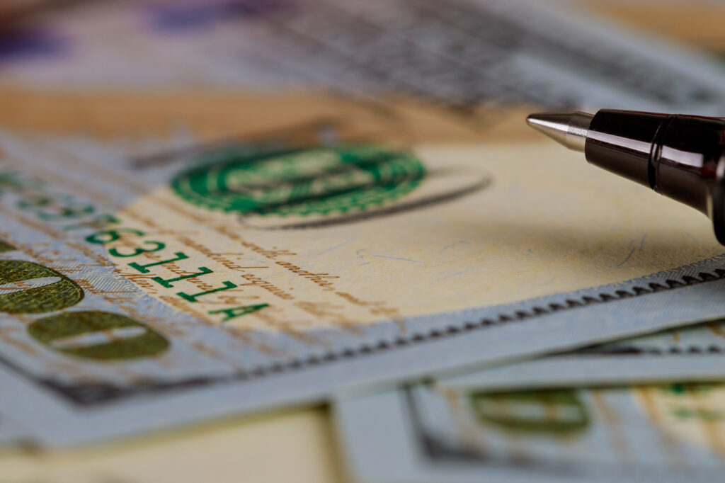 Close-up of a fountain pen nib resting on a stack of banknotes, with a green seal visible in the foreground.