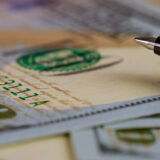 Close-up of a fountain pen nib resting on a stack of banknotes, with a green seal visible in the foreground.
