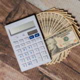 White calculator with blue buttons next to a fan of one-dollar bills on a wooden surface