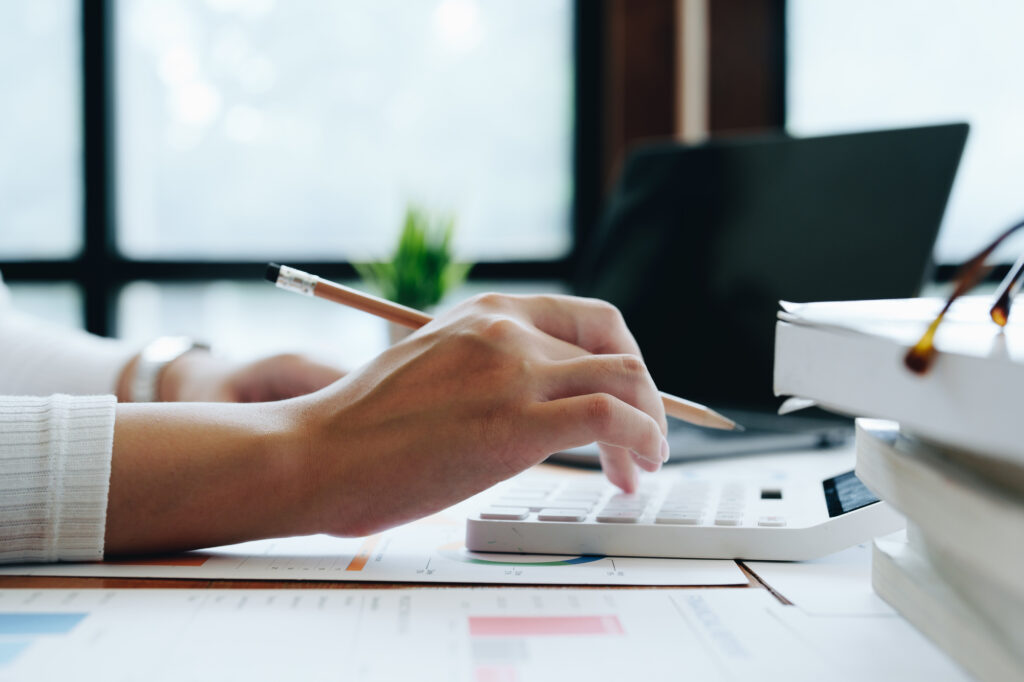 Hands writing with a pencil on financial documents and a calculator on a bright office desk with charts in view.