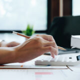 Hands writing with a pencil on financial documents and a calculator on a bright office desk with charts in view.