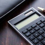 Close-up of a gray calculator on a dark wooden desk, with a brown-and-gold pen nearby